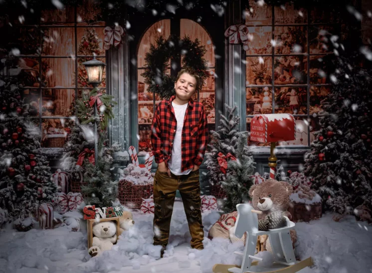 Un garçon vêtu d'une chemise à carreaux rouges et d'un pantalon kaki sourit dans une scène de Noël enneigée et festive en plein air, entouré d'arbres décorés, de cadeaux, de peluches et de chutes de neige. Cette photo a été prise par Photographe Iris Seine-et-Marne (77).