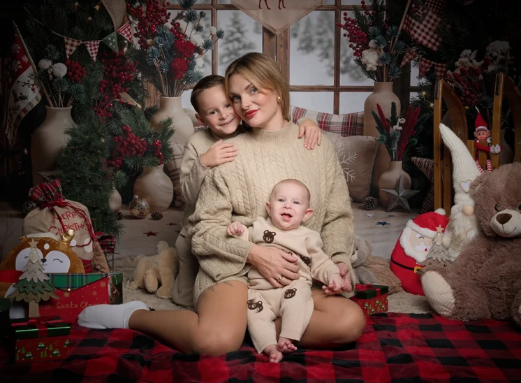 Une femme est assise sur une couverture à carreaux avec deux jeunes enfants, entourée de décorations de Noël et de cadeaux. Cette scène a été immortalisée par la photographe Iris Seine-et-Marne (77) dans une pièce chaleureuse et festive où trône un grand ours en peluche.