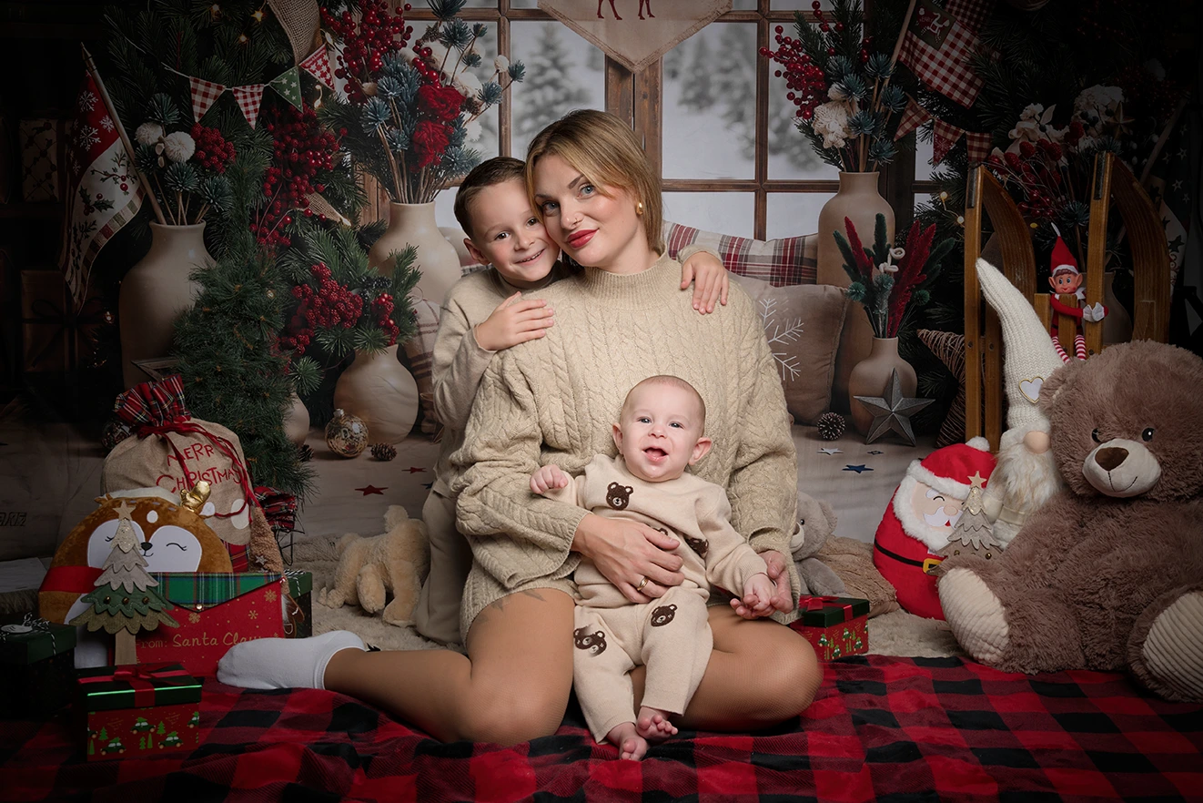 Une femme est assise sur une couverture à carreaux avec deux jeunes enfants, entourée de décorations de Noël et de cadeaux. Cette scène a été immortalisée par la photographe Iris Seine-et-Marne (77) dans une pièce chaleureuse et festive où trône un grand ours en peluche.