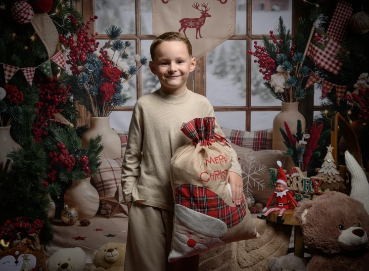 Un petit garçon souriant, vêtu d'un pyjama crème, tient un sac-cadeau à carreaux portant l'inscription Merry Christmas ; il se tient debout dans une pièce décorée pour les fêtes, photographié par Iris Seine-et-Marne (77). En arrière-plan, on aperçoit des fenêtres recouvertes de neige.
